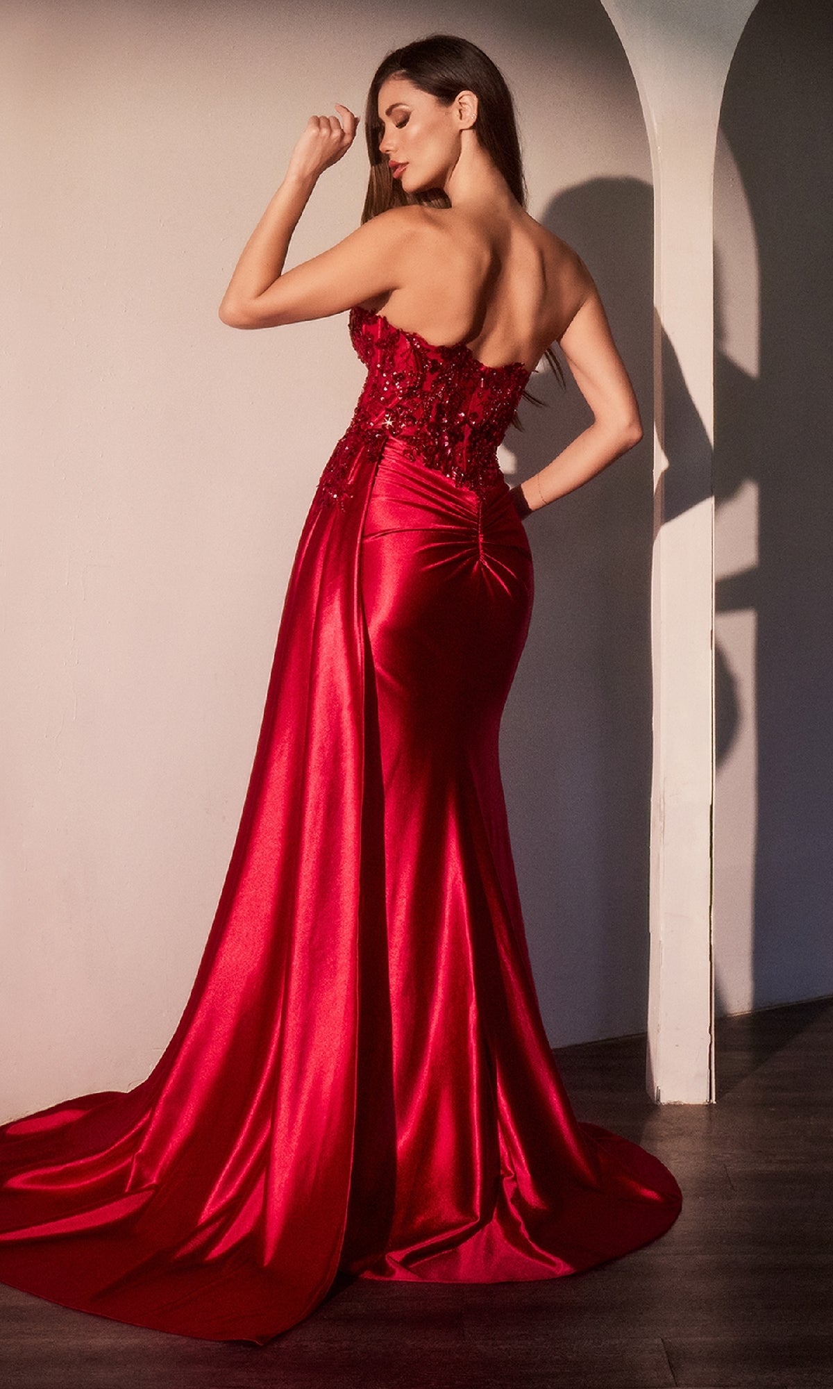 A woman stands with her back to the camera in the Ladivine CDS530 Lace-Corset Strapless Prom Gown, a floor-length red satin dress with a fitted bodice and flowing train that radiates elegance.